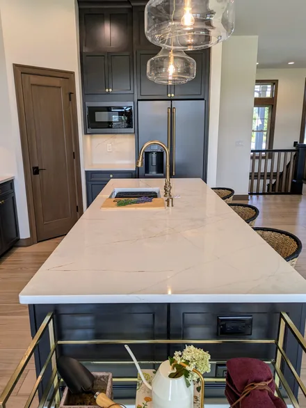 Long white quartz kitchen island with a brass faucet, seen from the end, with dark cabinets behind it